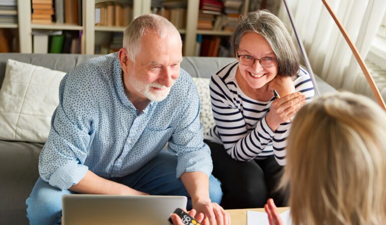 Older man and woman sit on a living room sofa, looking at a laptop as they discuss with a person off screen.