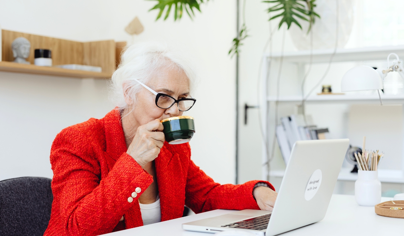 Older woman with white hair and glasses sipping from a dark green mug while working on a silver laptop at a white desk in a bright room.