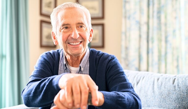 Smiling elderly man sitting on a blue sofa in a bright living room.