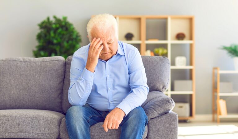 Older man sits on a gray couch with one hand pressed to his forehead, looking distressed at home.